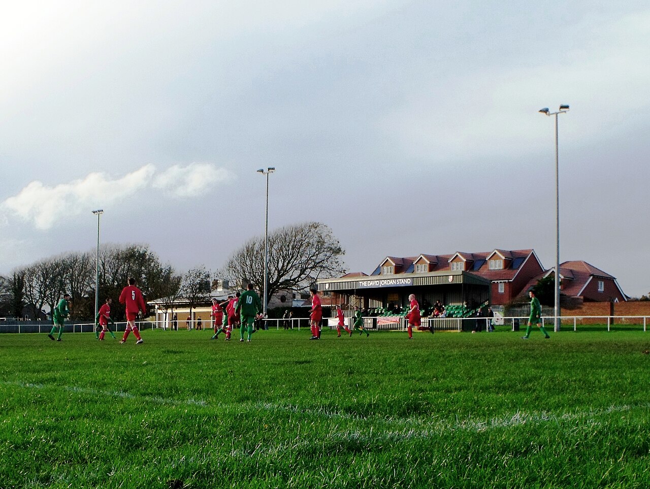 A grassroots match under a moody sky, spectators in a small wooden stand